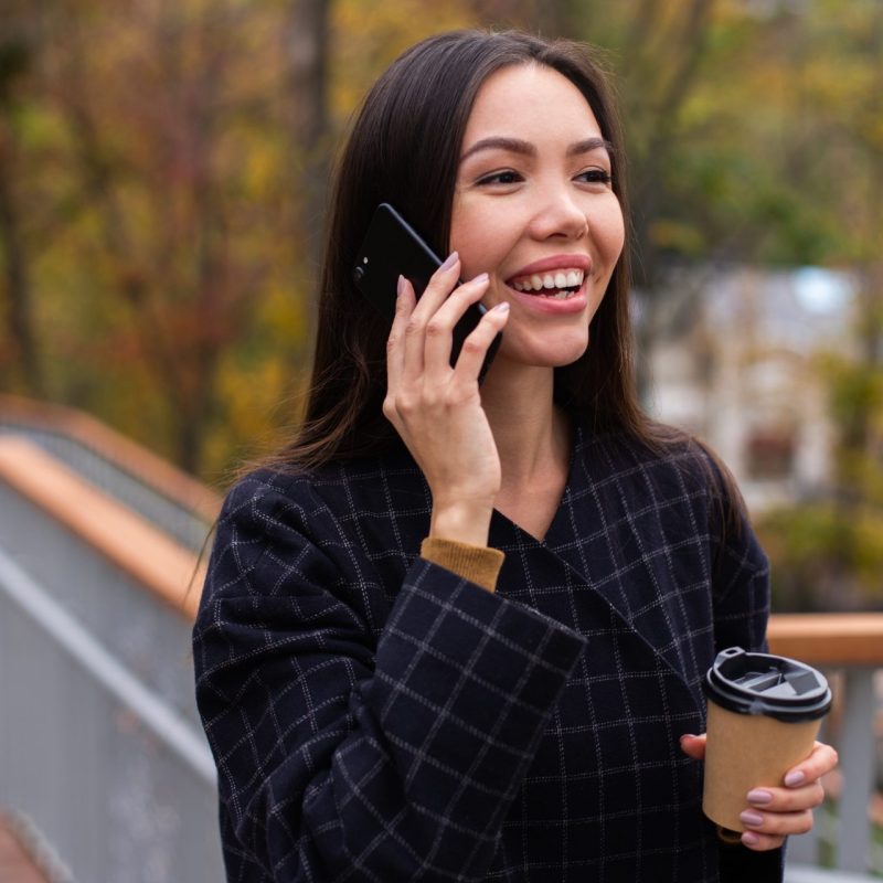 young positive casual woman in coat with coffee while on a call dialing a 0345 number in UK