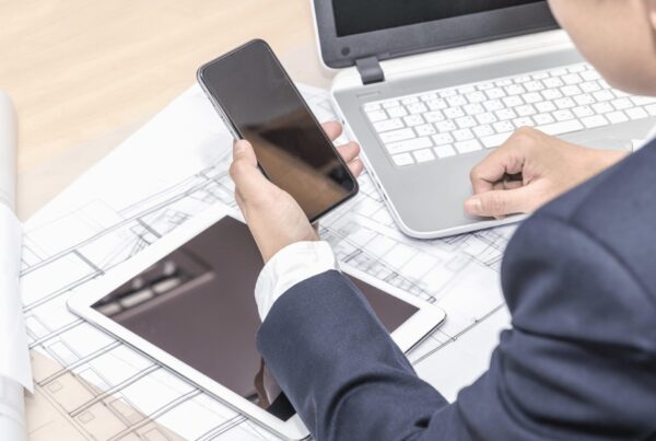 A man in a business suit working at a desk with a laptop, smartphone, and tablet placed over architectural blueprints, suggesting multitasking in a design or planning workflow.