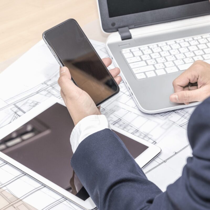 A man in a business suit working at a desk with a laptop, smartphone, and tablet placed over architectural blueprints, suggesting multitasking in a design or planning workflow.