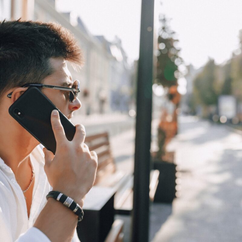 A man with dark hair and sunglasses, wearing a white shirt and a black bracelet, is seen in profile talking on a black smartphone. He appears to be calling an 0800 number on Tesco Mobile. The background shows a blurry outdoor street scene with trees and buildings, suggesting he is outside on a sunny day.