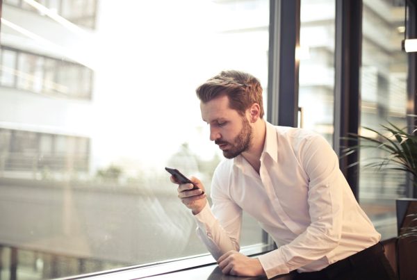 A person dressed in a white button-down shirt and black pants leans casually against a window, holding a smartphone and looking at the screen. Behind them, a blurred cityscape peeks through the glass, while a nearby plant adds a touch of greenery to the indoor setting. The moment captures a quiet pause in a modern, professional atmosphere.
