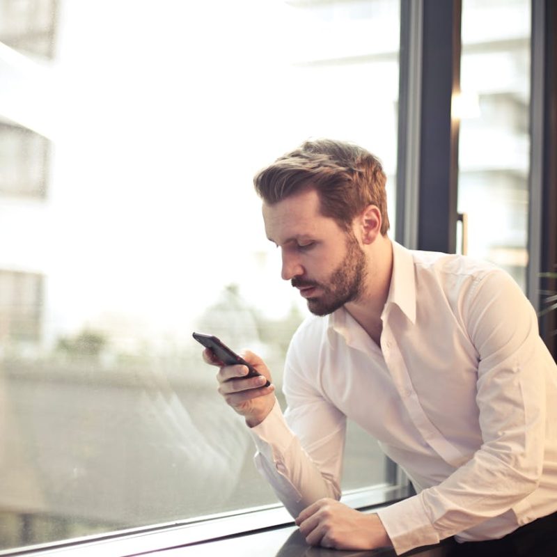 A person dressed in a white button-down shirt and black pants leans casually against a window, holding a smartphone and looking at the screen. Behind them, a blurred cityscape peeks through the glass, while a nearby plant adds a touch of greenery to the indoor setting. The moment captures a quiet pause in a modern, professional atmosphere.