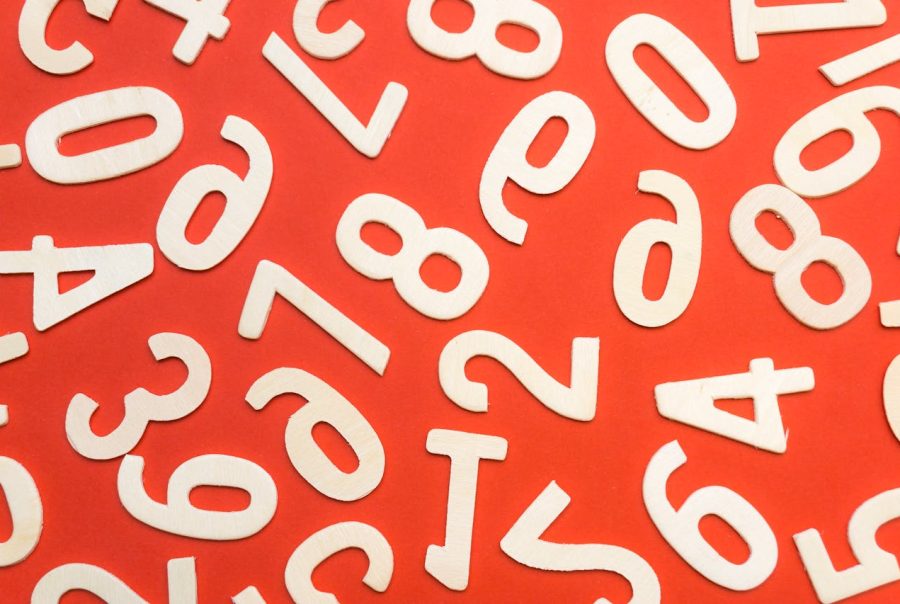 A cluster of white wooden number cutouts (including 2, 7, 8, and 9) arranged randomly on a bold red background. The light-colored wood contrasts sharply against the saturated red surface, creating an eye-catching, playful composition with a crisp, modern aesthetic.