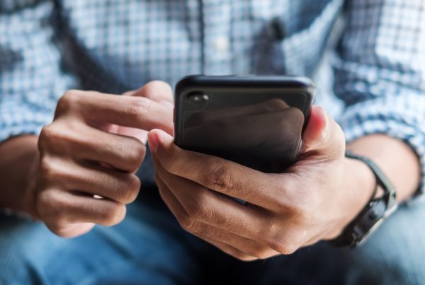 Close-up of a person wearing a blue and white checkered shirt and blue jeans, using a smartphone. The left wrist is adorned with a watch, while both hands are engaged with the phone screen, suggesting interaction or navigation. The setting is casual and focused on mobile use