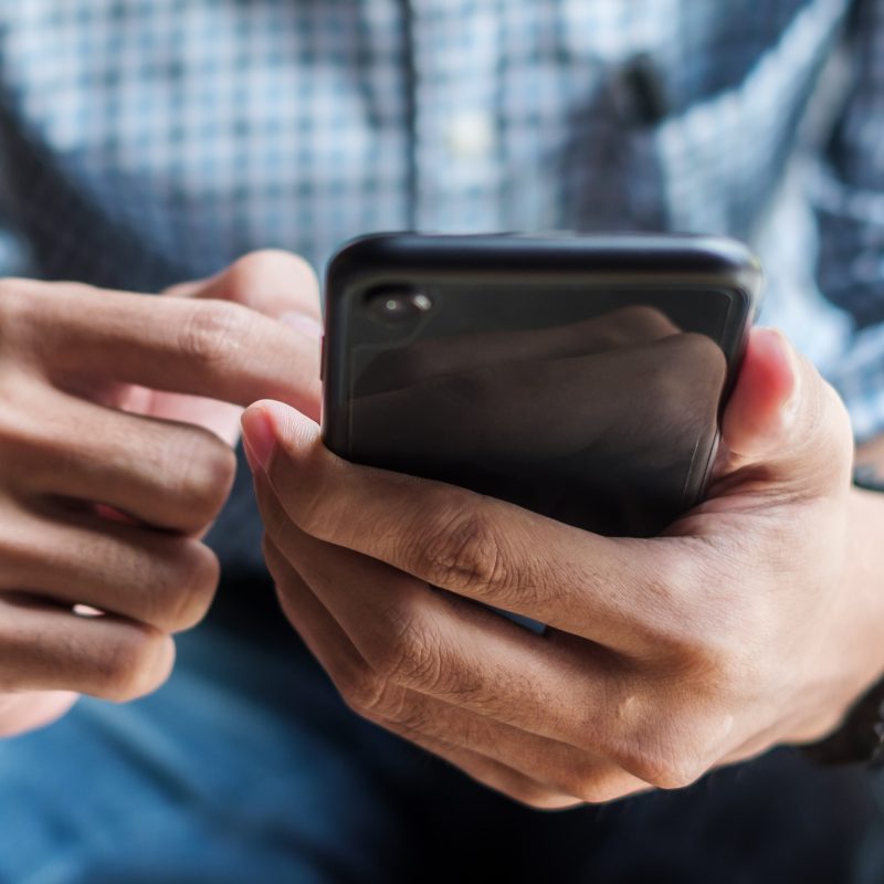 Close-up of a person wearing a blue and white checkered shirt and blue jeans, using a smartphone. The left wrist is adorned with a watch, while both hands are engaged with the phone screen, suggesting interaction or navigation. The setting is casual and focused on mobile use