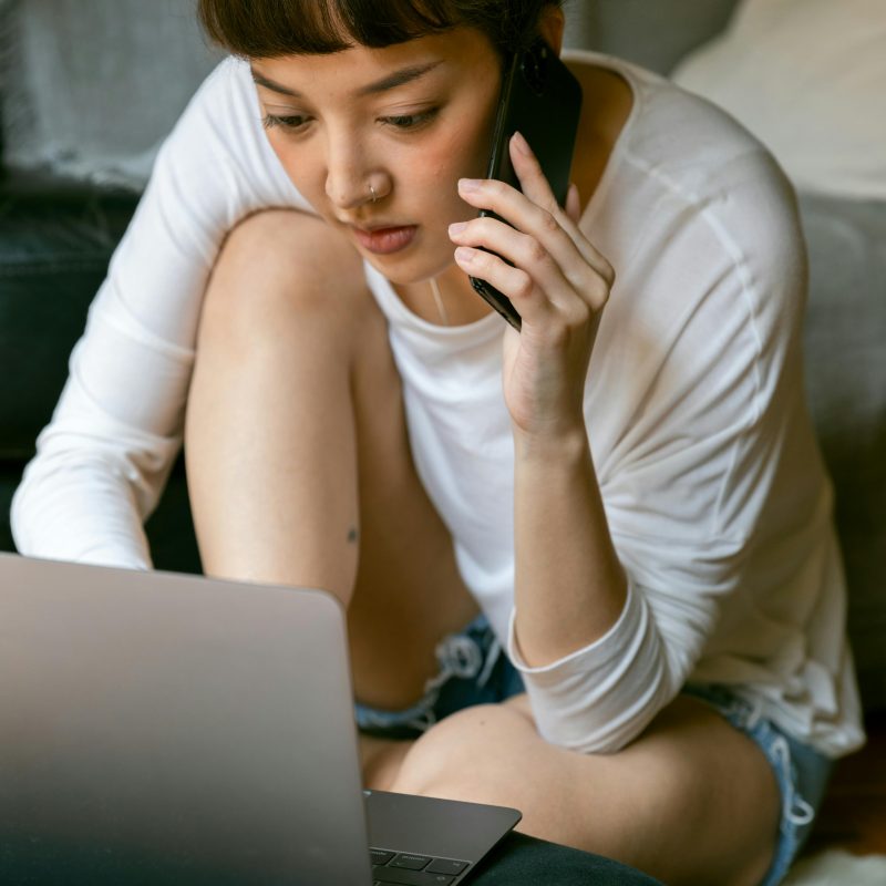 Casual home setup with a person lounging on a couch or bed, multitasking between a smartphone call and typing on a laptop. They're dressed in a white long-sleeve shirt and denim shorts, surrounded by soft furnishings like a pillow and blanket that create a relaxed atmosphere.