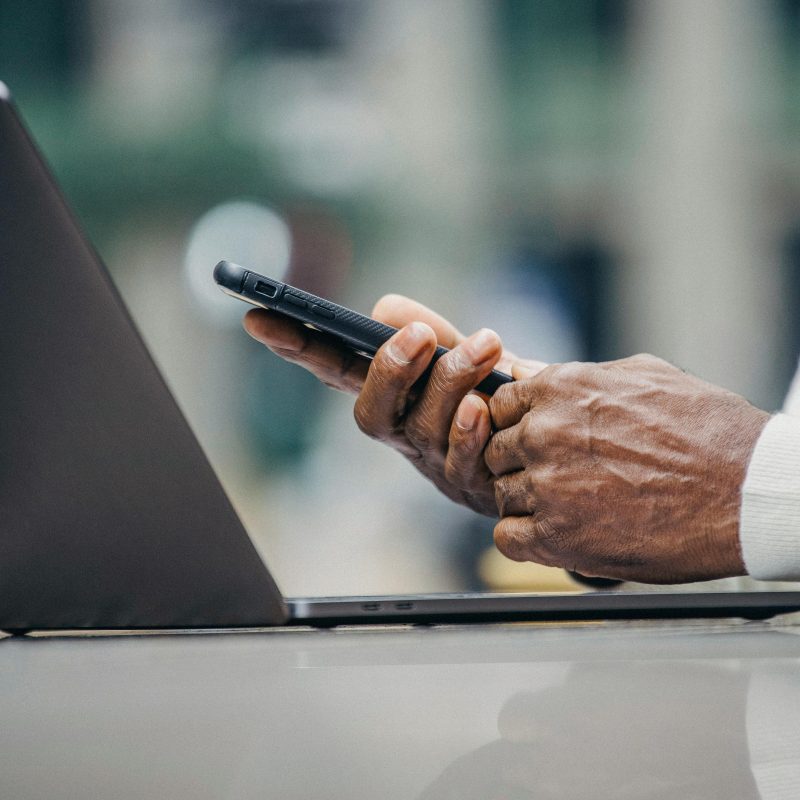 Close-up shot of a person wearing a light-colored long-sleeve shirt, holding a smartphone with both hands while seated in front of an open laptop. The focus is on the hands, phone, and laptop, with a softly blurred background suggesting a tech-savvy workspace or remote work scenario.