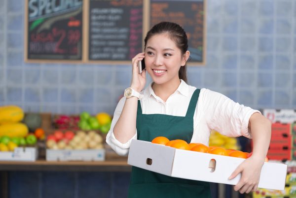 The image shows a young woman smiling as she holds a box filled with oranges in one hand, while talking on her phone with the other hand. She is dressed in a white shirt and a green apron, standing in what looks like a grocery store or market with colorful fruits in the background.