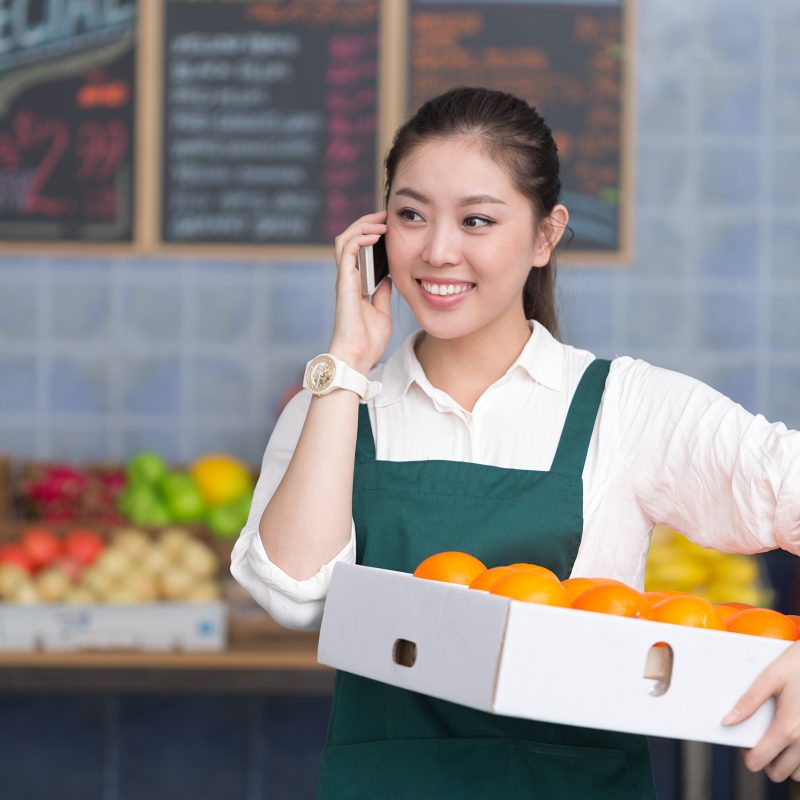 The image shows a young woman smiling as she holds a box filled with oranges in one hand, while talking on her phone with the other hand. She is dressed in a white shirt and a green apron, standing in what looks like a grocery store or market with colorful fruits in the background.
