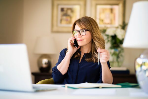 This image depicts a woman with glasses speaking on the phone. She appears to be sitting at a desk with a laptop in front of her and a pen in her hand. The setting is cozy and professional, with soft lighting and a minimalist background featuring plants and framed artwork on the walls.