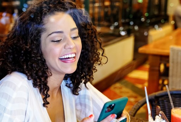 Smiling woman with curly hair sitting at a restaurant table, looking at her smartphone while laughing, with a colorful drink and utensils in front of her.