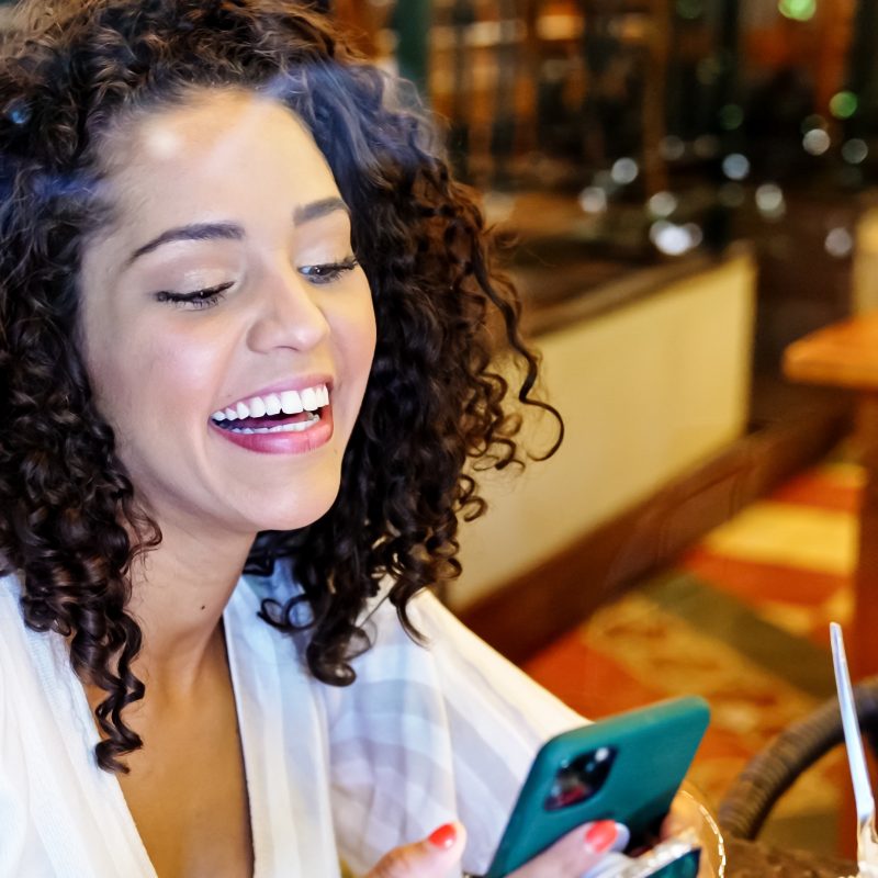Smiling woman with curly hair sitting at a restaurant table, looking at her smartphone while laughing, with a colorful drink and utensils in front of her.