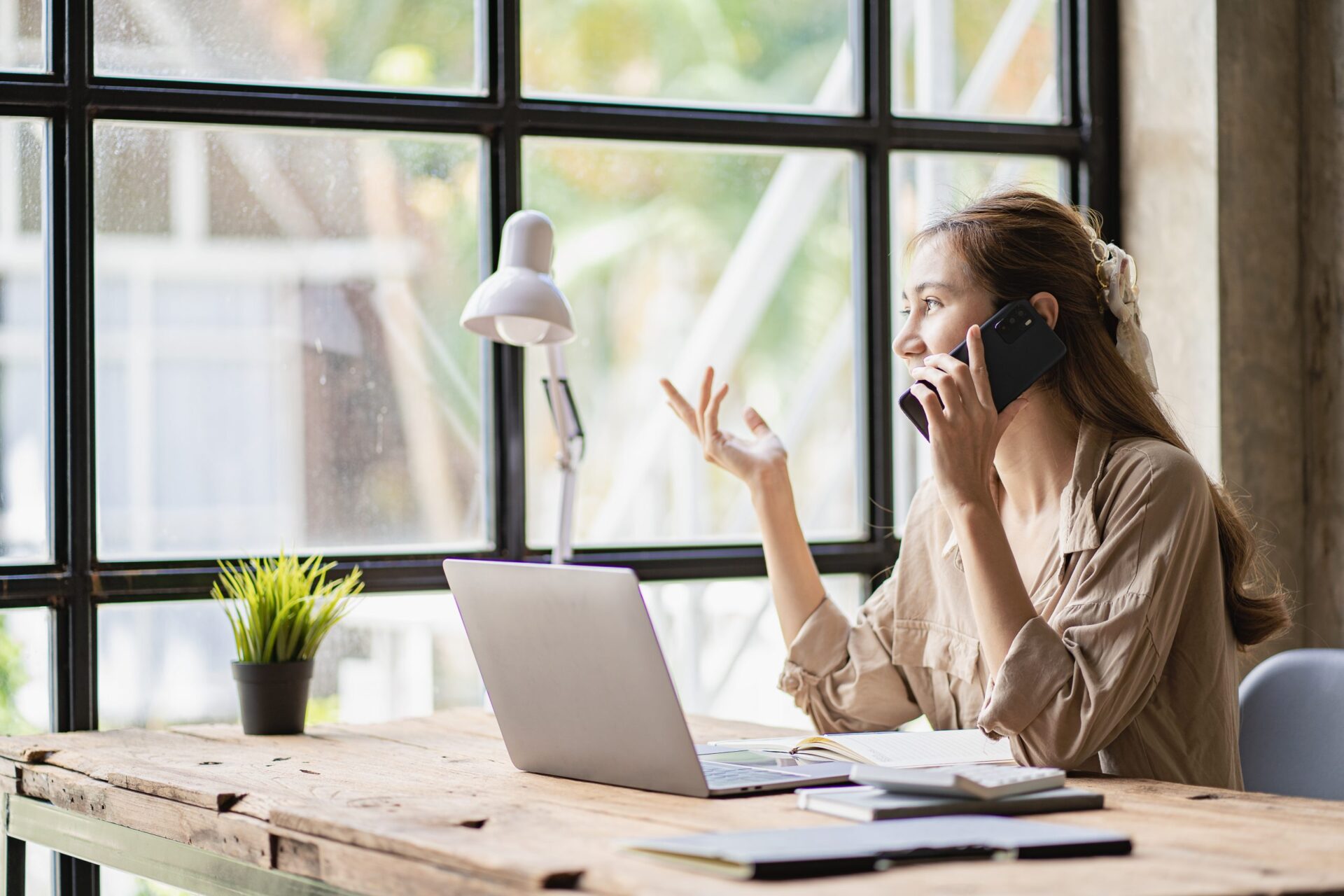 Young woman sitting at a wooden desk in front of a large window, talking on the phone while gesturing with her hand, with a laptop, notebook, and desk lamp in front of her.