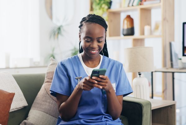 Smiling nurse in blue scrubs sitting on a couch and looking at her smartphone, in a cozy and well-lit living room with a desk and computer in the background.