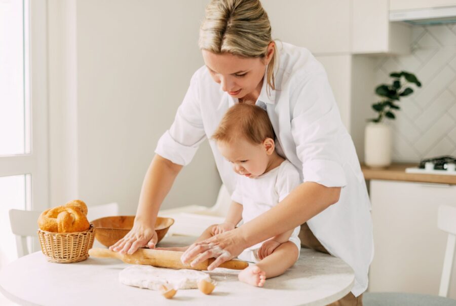 Adult and child baking together in a cozy kitchen, rolling out dough on a table scattered with flour, eggshells, and bread.