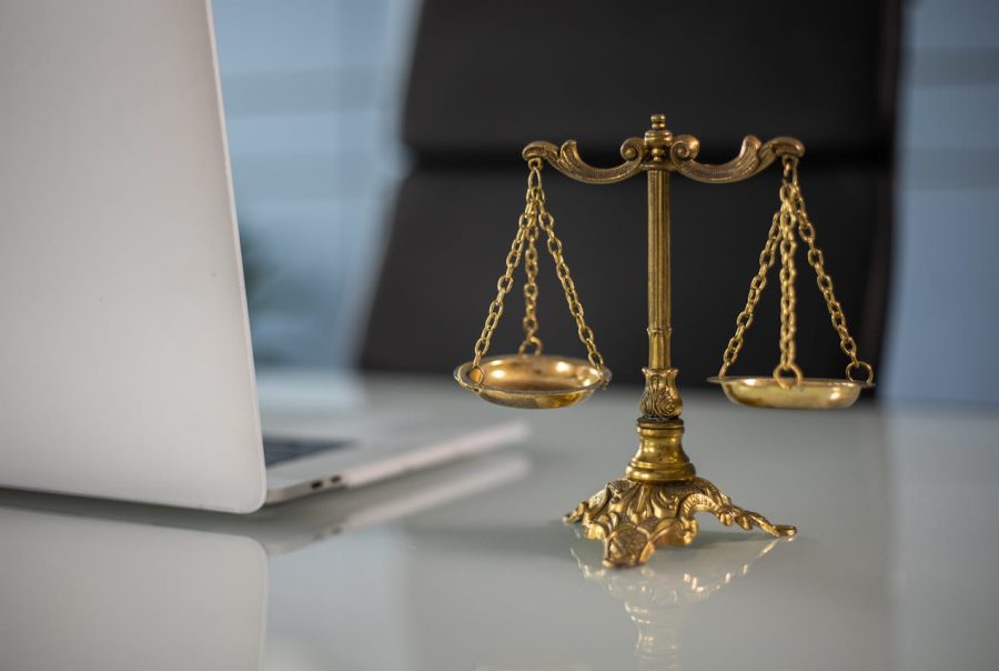 The image shows a golden scale of justice placed on a glass desk next to a laptop. The background is blurred, but it suggests a modern office setting. The scale symbolizes justice, and the laptop indicates a professional, possibly legal, work environment.
