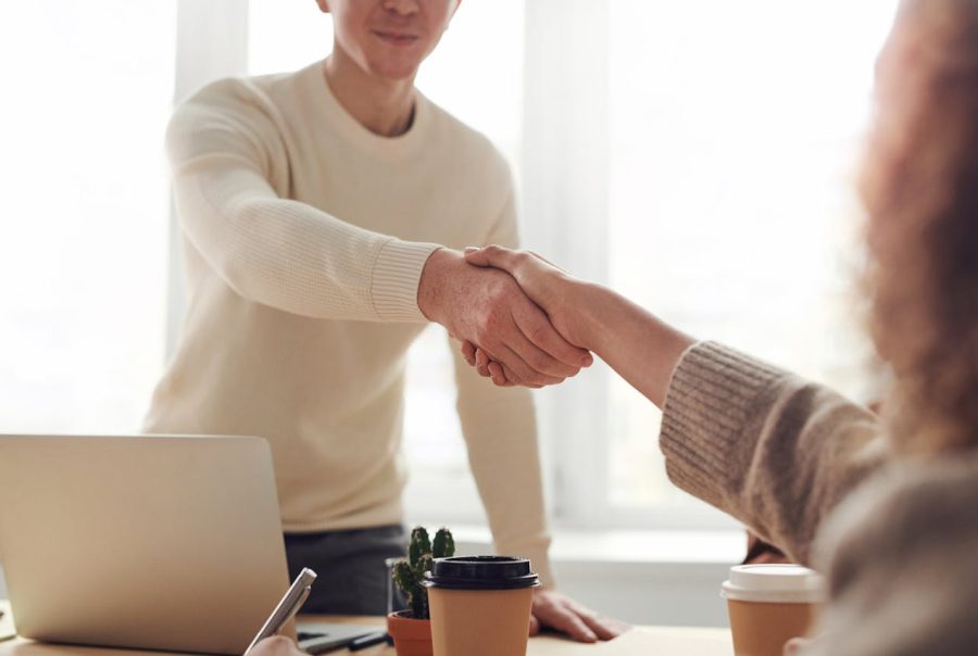 Two individuals are shaking hands in a bright, modern office space. One person, wearing a light-colored sweater, is extending their hand towards the other, while a warm smile is visible on their face. A coffee cup and a small potted cactus are placed on the table in the foreground, and a laptop is partially visible. The background features soft, natural light coming through large windows.