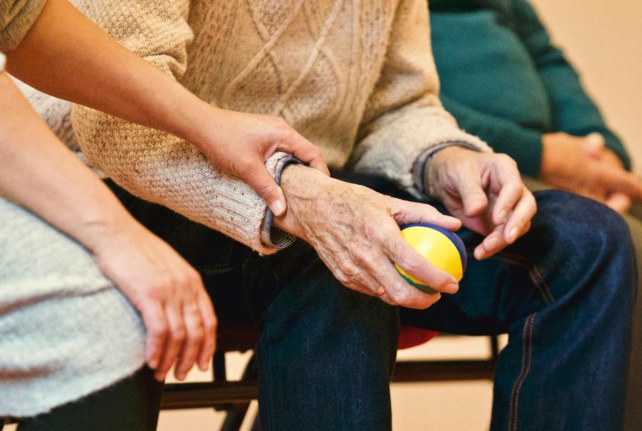 An elderly person is holding a small, colorful stress ball while another person's hand is gently placed on their arm. The image focuses on the close-up interaction, with the elderly person wearing a knitted sweater and sitting in a group of individuals. The warm, caring touch of the second person suggests a moment of comfort or support.